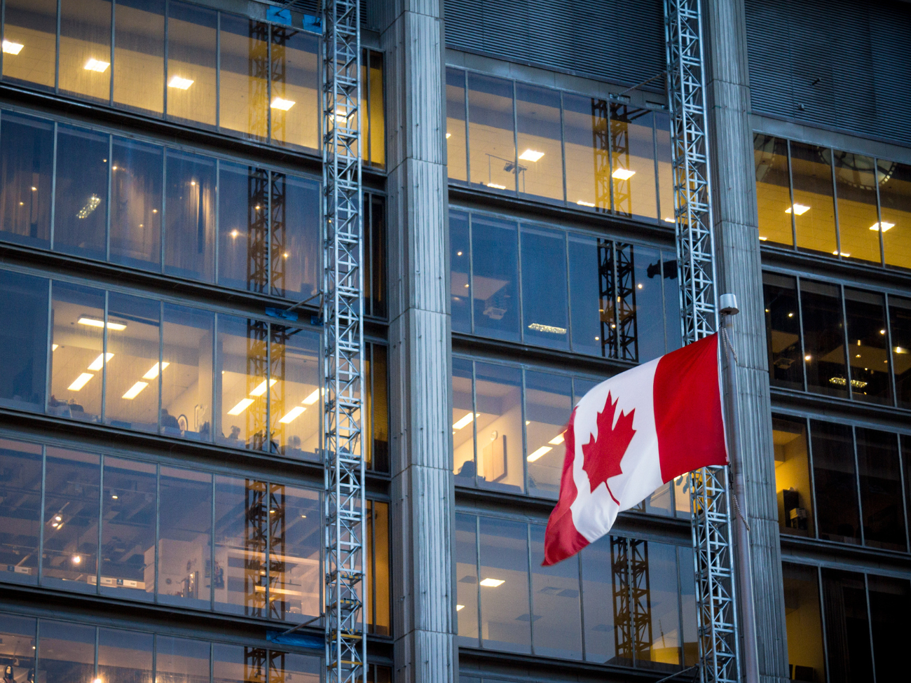 Canadian flag waving in front of a modern glass office building during the evening.